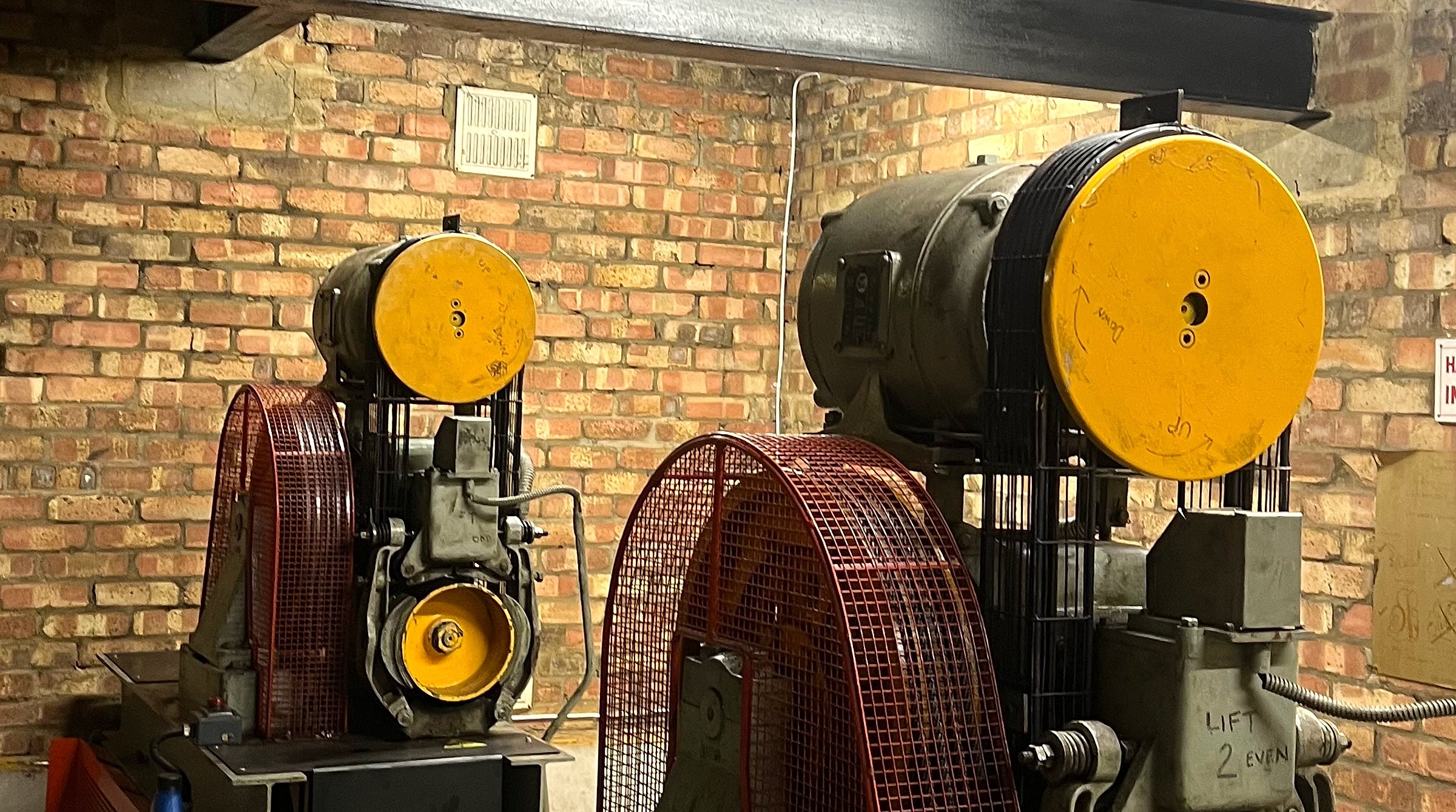 Two ageing geared traction lift machines in a brick-walled machine room